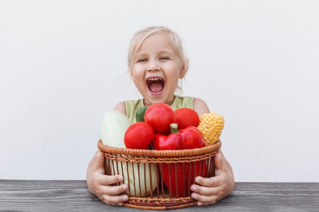 Mała dziewczynka siedząca przy stole przytula kosz pełen świeżych warzyw i raduje się