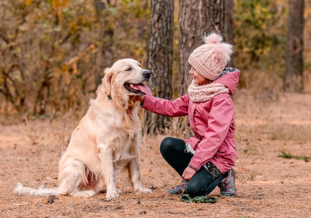 Mała dziewczynka siedzi bokiem, trzymając w rękach smycz golden retrievera w jesiennym lesie