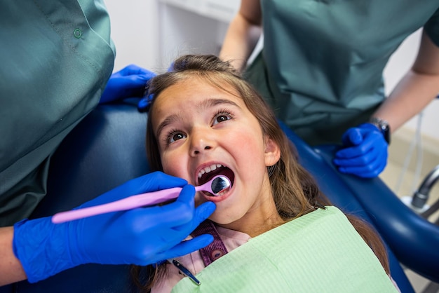 Mała Dziewczynka Siedzi Na Fotelu Dentystycznym I Dwie Dentystki Leczą Zęby Dziecka Dwie Dentystki Zabawiają Małego Pacjenta