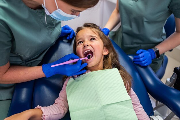 Mała dziewczynka siedzi na fotelu dentystycznym i dwie dentystki leczą zęby dziecka