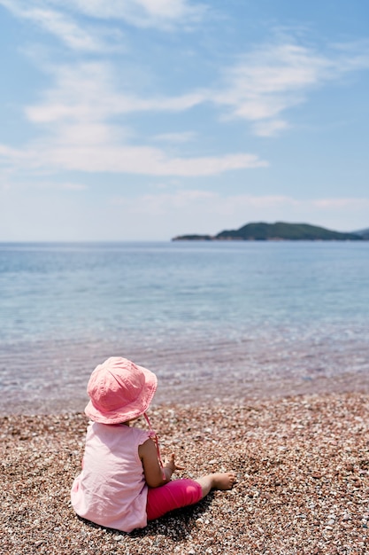 Mała dziewczynka siedzi na kamienistej plaży w pobliżu widoku wody z tyłu