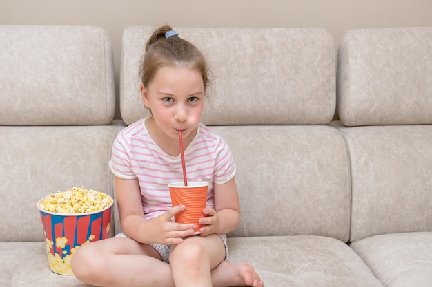 mała dziewczynka siedzi na kanapie w domu pije koktajl ze słomki, obok jest wielka szklanka popcornu