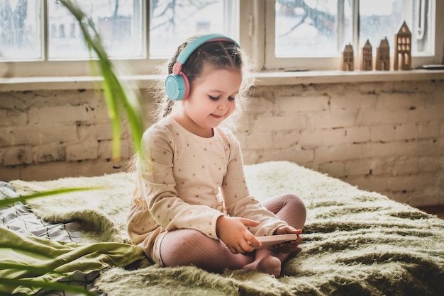 Mała dziewczynka siedzi na łóżeczku w słuchawkach dziecko lekkomyślnie gra w gry i słucha muzyki