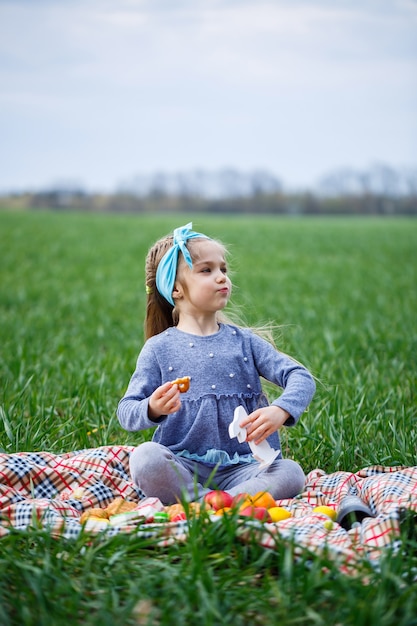 Mała dziewczynka siedzi na narzucie i je ciasteczka i marmoladę, zielona trawa na polu, słoneczna wiosenna pogoda, uśmiech i radość dziecka, błękitne niebo z chmurami