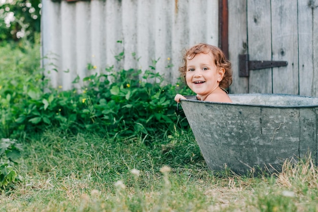 Mała dziewczynka siedzi w metalowej wannie w zielonym ogrodzie w upalny letni dzień ząbkowany uśmiech spójrz na aparat