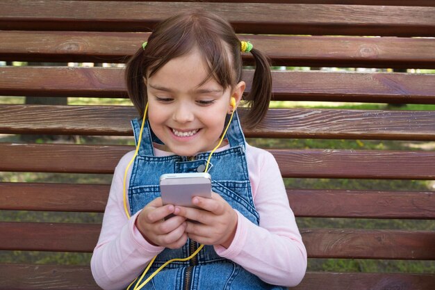 Mała dziewczynka słucha muzyki w słuchawkach telefonem i uśmiecha się w parku na ławce