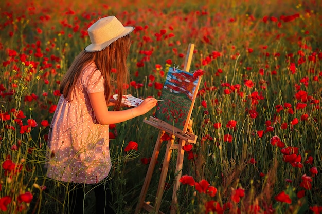 Zdjęcie mała dziewczynka stoi na polu czerwonych maków o zachodzie słońca i maluje na płótnie umieszczonym na stojaku do rysowania