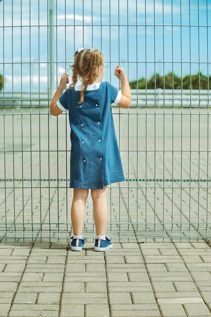mała dziewczynka stoi przy barierce z wysokiego siatkowego ogrodzenia i trzyma się jej rękami