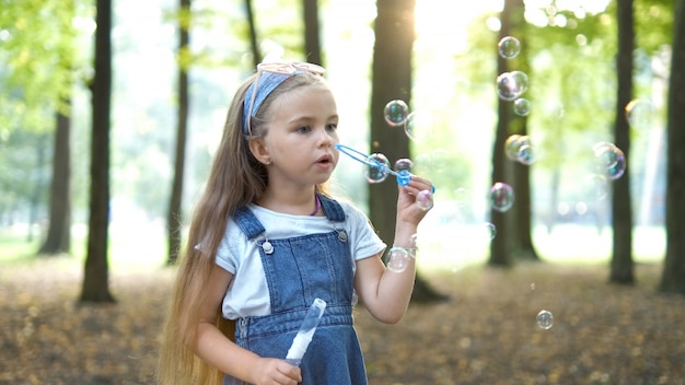 Mała dziewczynka szczęśliwe dziecko dmuchanie baniek mydlanych na zewnątrz w zielonym parku. Koncepcja działalności na świeżym powietrzu latem.