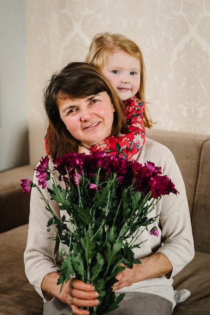 Zdjęcie mała dziewczynka trzyma bukiet kwiatów robi niespodziankę dla babci gratulującej jej urodzin szczęśliwa babcia i wnuczka obejmują się razem w domu dzień kobiet