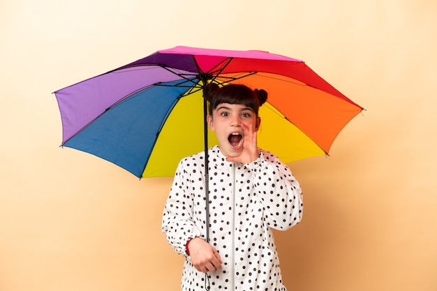 Mała dziewczynka trzyma parasol na białym tle na beżowej ścianie z zaskoczeniem i zszokowanym wyrazem twarzy