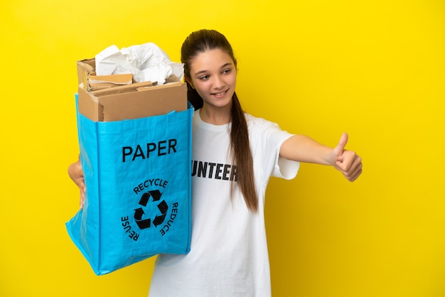 Mała dziewczynka trzyma torbę do recyklingu pełną papieru do recyklingu na izolowanym żółtym tle, pokazując gest kciuka w górę