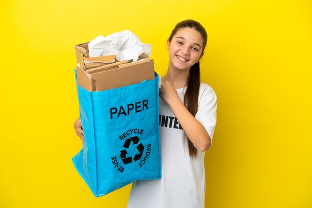 Mała dziewczynka trzyma torbę do recyklingu pełną papieru do recyklingu na izolowanym żółtym tle świętującym zwycięstwo