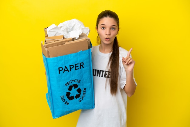 Mała dziewczynka trzyma torbę do recyklingu pełną papieru do recyklingu na na białym tle żółtym, zamierzając zrealizować rozwiązanie, podnosząc palec w górę