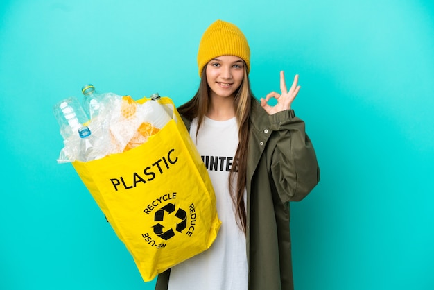 Mała dziewczynka trzyma torbę pełną plastikowych butelek do recyklingu na odosobnionym niebieskim tle pokazującym znak ok palcami