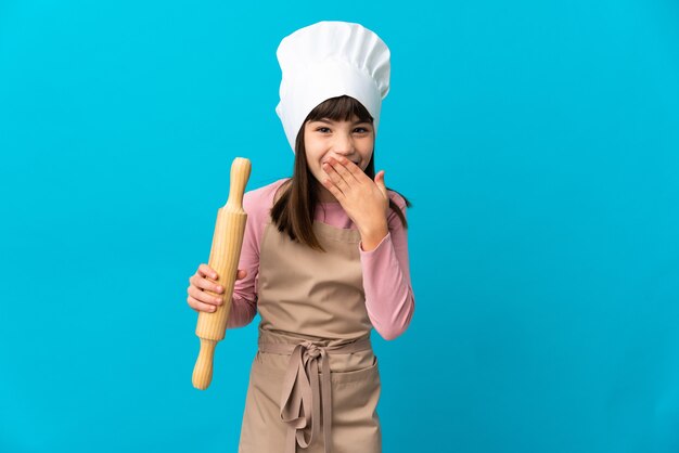 Mała dziewczynka trzyma wałek do ciasta na białym tle na niebieskiej ścianie szczęśliwa i uśmiechnięta obejmująca usta ręką