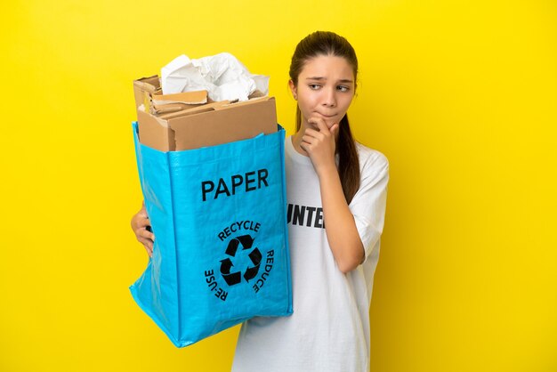 Mała dziewczynka trzymająca torbę do recyklingu pełną papieru do recyklingu na izolowanym żółtym tle, mająca wątpliwości i myśląca