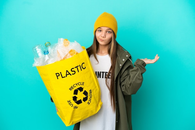 Mała dziewczynka trzymająca torbę pełną plastikowych butelek do recyklingu na odosobnionym niebieskim tle, mająca wątpliwości podczas podnoszenia rąk