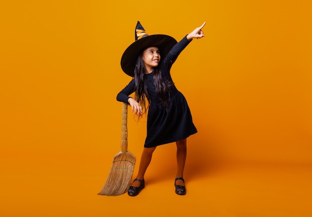 Zdjęcie mała dziewczynka ubrana jak czarownica halloween w czarnej sukience i kapeluszu leci na miotle na żółtym tle.