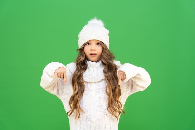 Mała dziewczynka ubrana w ciepły sweter i zimową czapkę z pięknymi kręconymi włosami na białym tle pokazuje na dole ogłoszenia Dziecko w ciepłych ubraniach w zimnych porach roku