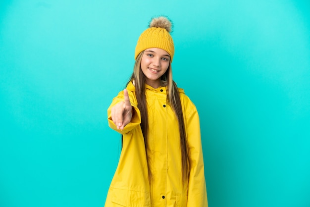 Mała dziewczynka ubrana w przeciwdeszczowy płaszcz na odosobnionym niebieskim tle pokazująca i unosząca palec