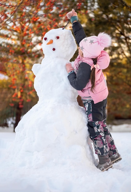 Mała dziewczynka ubrana w zimowe ubrania robiące włosy z gałązkami dla bałwana w parku w ciągu dnia podczas spaceru z drzewami i jasnym światłem słonecznym w tle Rodzice spędzają czas z dziećmi