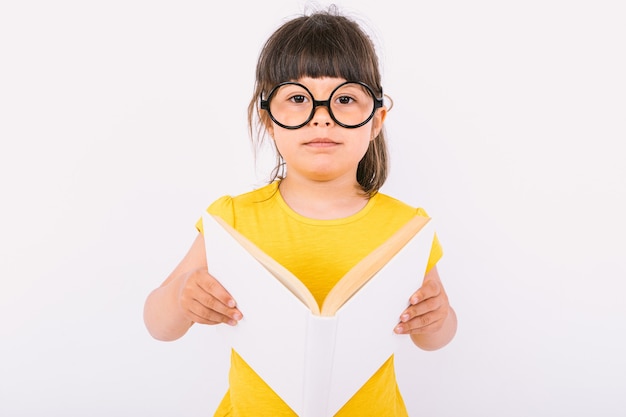 Mała dziewczynka ubrana w żółtą koszulkę i okrągłe czarne okulary, trzymająca w dłoniach otwartą książkę na białym tle