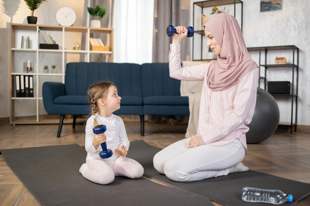 Mała dziewczynka uprawia sport ze swoją muzułmańską matką siedzącą na macie i wykonującą ćwiczenia z hantlami