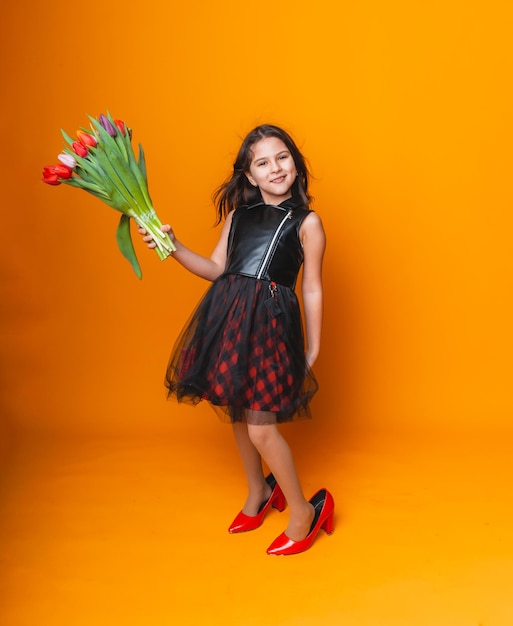 Mała dziewczynka uśmiecha się w sukience i dużych czerwonych butach trzyma bukiet kwiatów na żółtym tle miejsca na tekst