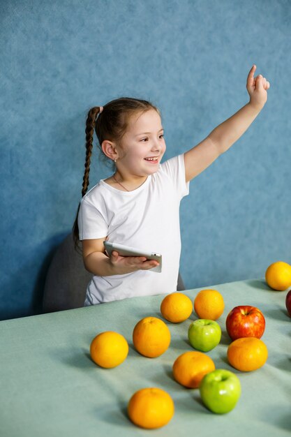 Mała dziewczynka w białej koszulce trzyma tablet w dłoniach i studiuje owoce