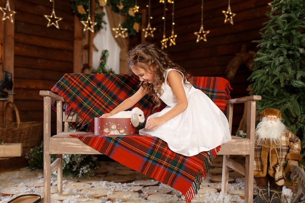 Mała dziewczynka w białej sukni siedzi na ławce i otwiera pudełko z białym królikiem. Magic Light in Night Xmas Tree Interior