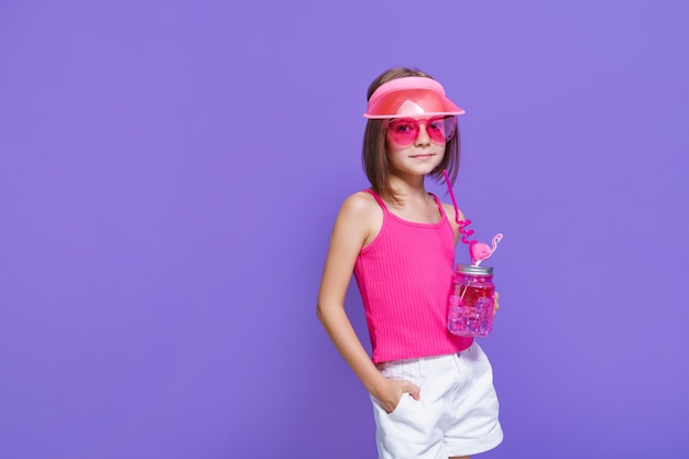 Mała dziewczynka w białych spodenkach, różowej koszulce, modnych okularach, z drinkiem w dłoni i letnim daszkiem