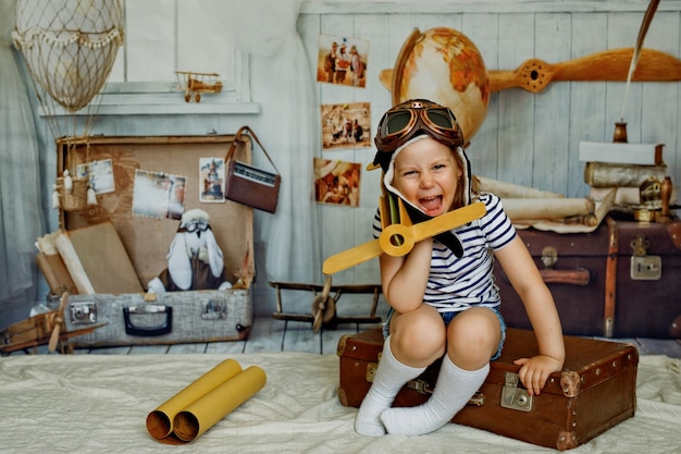 Mała Dziewczynka W Czapce Siedzi Na Walizce W Stylu Retro I Trzyma W Ręku Drewniany Samolot