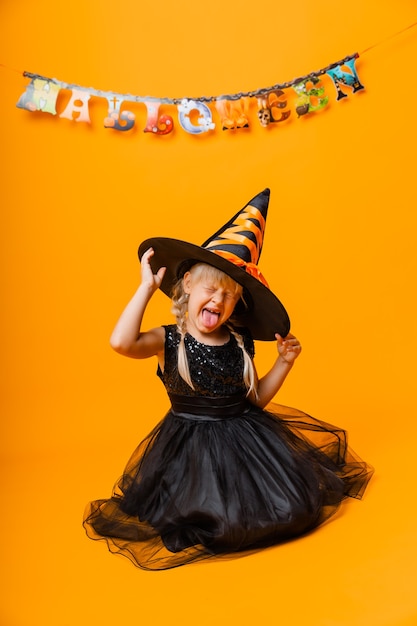 Mała dziewczynka w czarnym kostiumie halloween, śmiejąc się i patrząc w kamerę, skacząc i bawiąc się, na białym tle na żółtym tle. Halloween