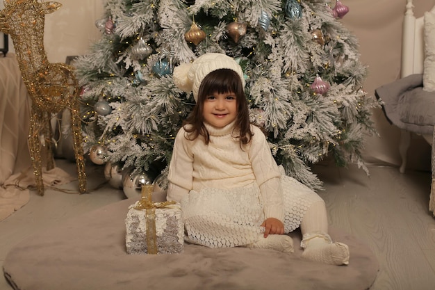 mała dziewczynka w dzianinowej czapce i białej sukience siedzi na choince obok pudełka z prezentami