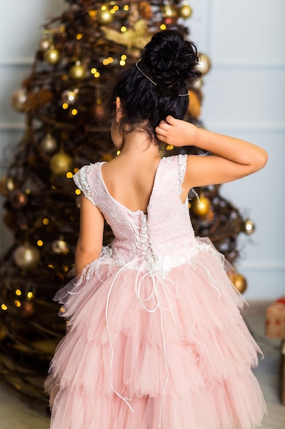 Mała Dziewczynka W Eleganckiej Różowej Sukience Stoi Tyłem Do Aparatu I Patrzy Na Prezenty Pod Drzewem