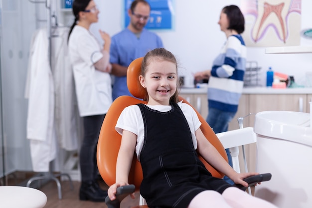 Mała dziewczynka w gabinecie dentystycznym, pokazując ząb mleczny, podczas gdy rodzic rozmawia z lekarzem dentystą. Dziecko z matką podczas badania zębów u stomatologa siedzącego na krześle.