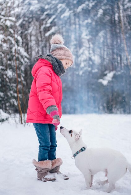 Mała dziewczynka w jasnej kurtce bawi się w zimowym, zaśnieżonym lesie ze swoim psem jack russell terrierem