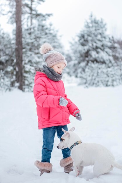 Mała dziewczynka w jasnej kurtce bawi się w zimowym, zaśnieżonym lesie ze swoim psem jack russell terrierem