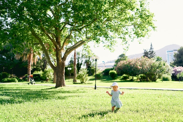 Mała dziewczynka w kapeluszu spaceruje po zielonym trawniku na tle drzew i kwitnących krzewów