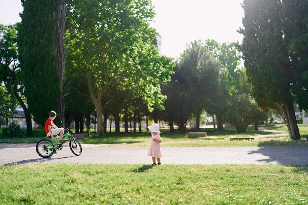 Mała dziewczynka w kapeluszu stoi na żwirowej ścieżce w parku i patrzy na rowerzystę