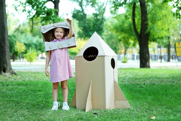 Mała dziewczynka w kasku pudełkowym stojąca w pobliżu kartonowej rakiety w parku