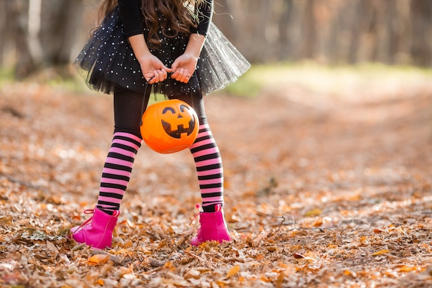 Zdjęcie mała dziewczynka w kostiumie wiedźmy trzyma na plecach wiadro z cukierkami w kształcie dyni na halloween