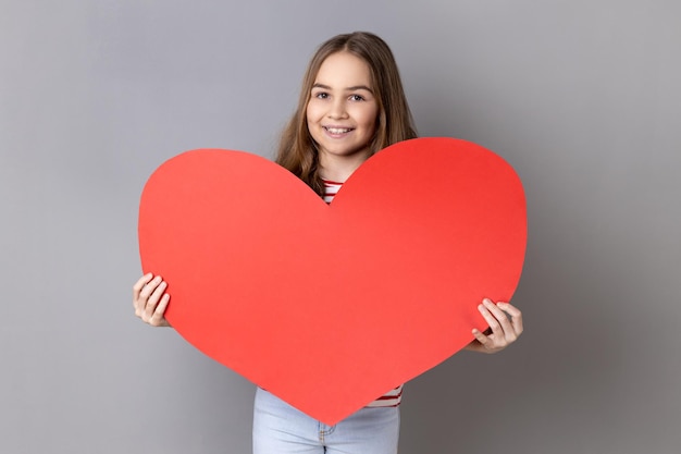 Mała dziewczynka w koszulce w paski trzymająca duże czerwone serce i uśmiechająca się do dziecka wyrażającego miłość