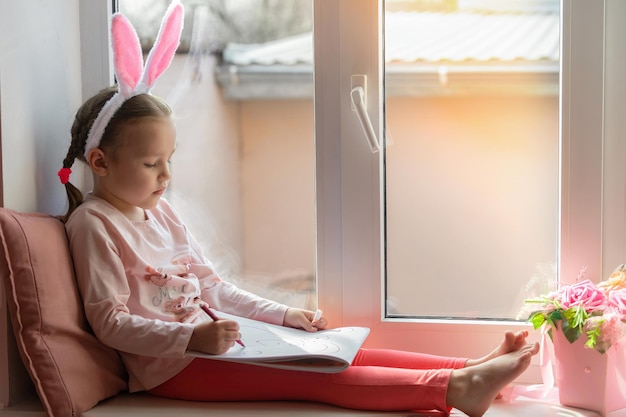 mała dziewczynka w króliczych uszach siedzi na oknie i rysuje króliczka w swoim albumie, przygotowując się do wielkanocy