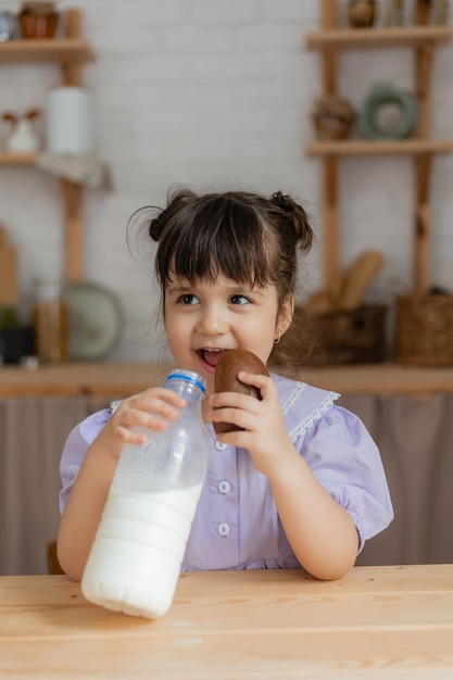 mała dziewczynka w liliowej sukience pije mleko i zjada bułkę przy kuchennym stole. miejsce na tekst, baner