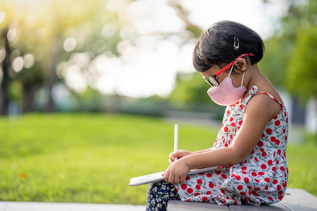 Mała dziewczynka w masce siedzi na trawniku i pisze w zeszycie.