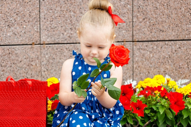 Mała dziewczynka w niebieskiej sukience i czerwonej kokardce siedzi z kolorowymi torebkami na ulicy obok kwiatów