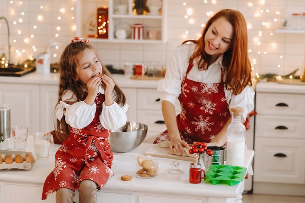 Mała dziewczynka w noworocznej kuchni siedzi na stole z ciasteczkami w dłoniach je je, a jej mama przygotowuje ciasto