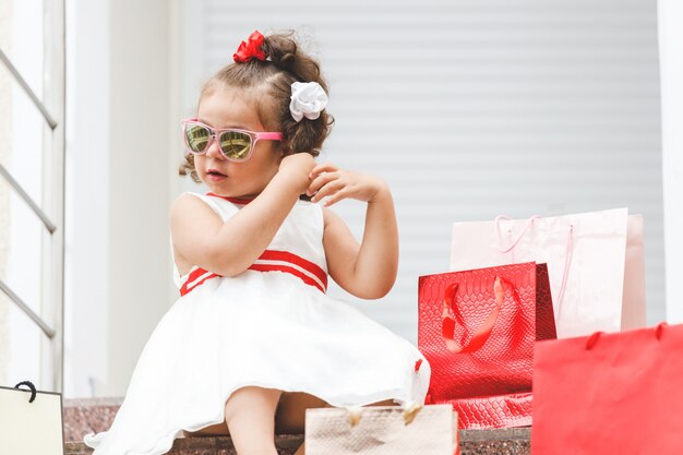 Mała dziewczynka w okularach przeciwsłonecznych siedzi na schodach w centrum handlowym z kolorowymi torbami